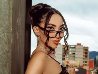 Stylish woman with glasses and a high bun posing on a balcony overlooking a city skyline at dusk.