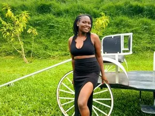 Smiling woman in a black crop top and slit skirt posing by a vintage carriage in a lush green field.