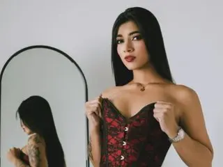 NashaFord poses in a red lace corset, looking at the camera with long dark hair against a plain white background.