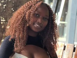 Nastyluna18 smiles while posing outdoors, showing her long, curly brown hair and cleavage in a black top.