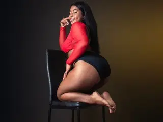 Smiling curvy woman in a red long-sleeve top and black shorts posing on a chair in a studio setting.