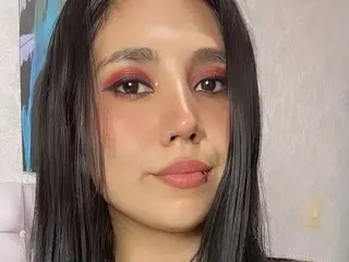 NatashaaHornny looks directly at the camera with long black hair and red eyeshadow, showing her face and shoulders against a plain background.
