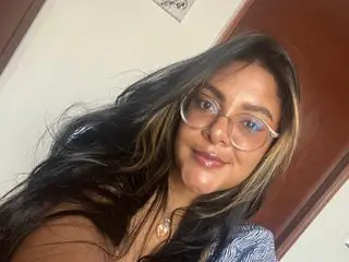 NathSaintss poses with long dark hair and glasses, wearing a blue patterned top against a plain white wall and dark door.
