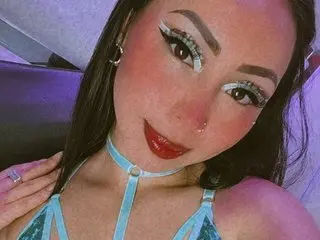 NathaliaPink poses with long dark hair and red lipstick, wearing a light blue strapset against a soft purple background.