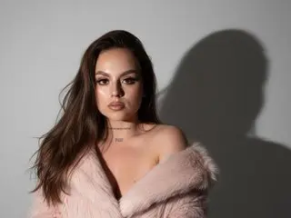 Natti_bluee poses with her long brown hair down, wearing a light pink furry shrug that leaves her chest and shoulders bare.