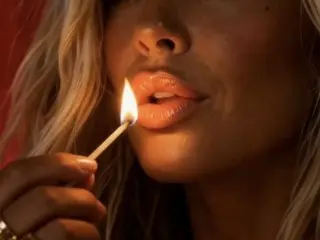 NaughtyLeahUK poses close up, holding a lit match near her open, wet lips and chin.