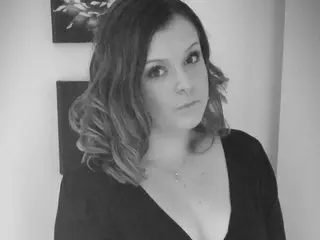NaughtyWifeUK poses with her wavy hair down, wearing a low-cut black top against a plain wall.