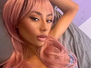 Naughty_Sapphire poses with her long pink hair, showing her face and shoulders against a light grey background.