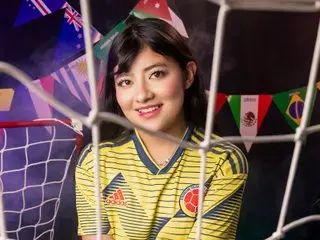 NekoPam poses in a yellow jersey with red and black trim, showing her face and shoulders against a dark background with colorful flags.