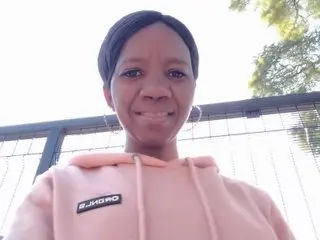 NewBootyXX — NewBootyXX smiles while wearing a light pink hooded sweatshirt against a bright outdoor background.