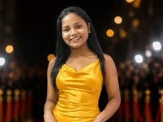 Nicki9693 smiles while wearing a yellow satin top, posing with her long dark hair down against a dark, blurry background.