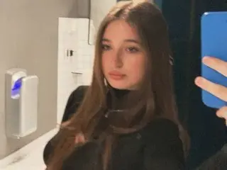 Nicole_De_Lux poses with long brown hair, wearing a black top against a neutral indoor background.