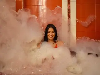 NicollColleman with long dark hair relaxing in a thick, white bubble bath, showing her bare shoulders and chest above the foam.