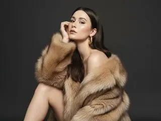 NikaHoney poses with her hand to her face, wearing a large, fluffy tan fur coat against a dark background.