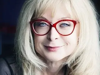 NinaHartley poses with a smile, showing her blonde hair and red-rimmed glasses against a dark background.