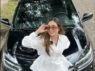 NiniThomson poses in a white long-sleeve shirt while sitting in the driver's seat of a black car, running her hand through her long brown hair.