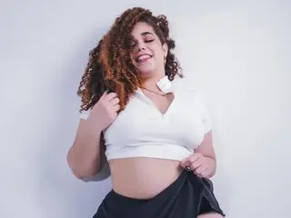 NinnaCurly smiles while posing in a white short-sleeve top and black skirt, showing her curly brown hair and midriff against a plain white background.