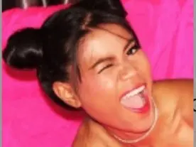 NizaThainy winks and smiles with her mouth open, showing her teeth against a bright pink background.