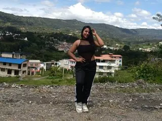 NoahWhyle poses outdoors in a black tank top and black pants, running her hand through her long dark hair against a natural landscape background.
