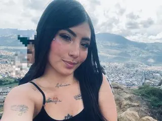 NoahdZoe poses with long dark hair and a light pink blush, showing her neck and chest tattoos against a soft outdoor background.