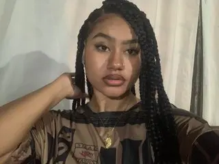 NoiraVale poses with her long dark braids, wearing a dark patterned top while looking directly at the camera.