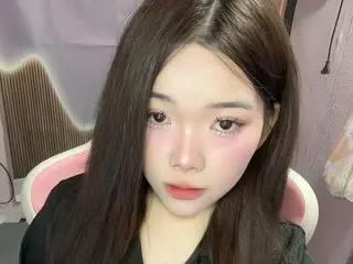 Nomi039 looks into the camera with long dark hair and pink makeup while sitting in a pink chair.