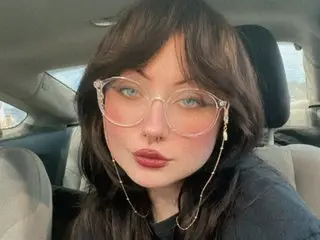 NovaMaelle18 poses close-up with long dark hair and glasses, wearing a dark top while sitting in a car seat.