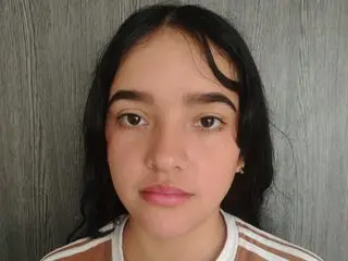 NoxuRose looks directly at the camera with long dark hair and a neutral expression, wearing a striped top against a grey wall.