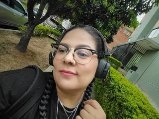 NyxxRaven wears black headphones and glasses while posing outdoors near green bushes and trees.