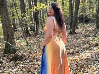 OLLIVYIA poses in a wooded setting, wearing a low-cut, orange and blue sundress that shows her cleavage and bare shoulders.