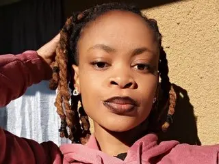 ObsessedMimi poses outdoors in a pink hoodie with her hair in long dreadlocks.