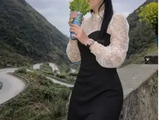 Oioine poses outdoors in a black sleeveless dress and a sheer white lace long-sleeve top, standing near a winding mountain road.