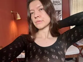 OliviaHawthorn poses with her hands behind her head, wearing a black long-sleeved patterned top with her long brown hair down.