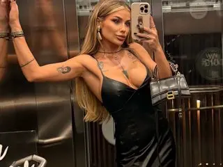 OliviaaBell poses in a mirror selfie wearing a black satin slip dress, showing her long blonde hair and cleavage in a dimly lit room.