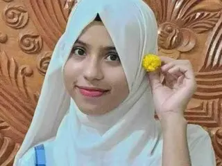 OliviaaIslam wearing a white hijab, looking at the camera with a soft smile against a brown carved wood background.