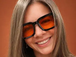 Oriana_thomson smiles while wearing large, orange-tinted sunglasses and letting her long, straight brown hair hang down.