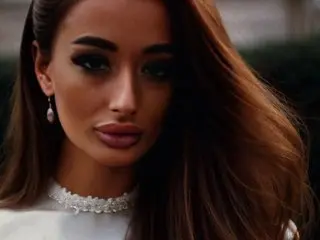 OutrageousOne poses with long brown hair and a direct gaze, wearing a white lace choker or collar against a soft, blurred background.
