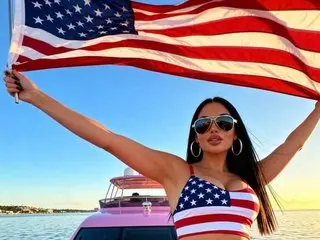 PalmBeachPrincesa poses outdoors by the water, wearing a red and white striped bikini top with her long dark hair down and arms spread wide behind a large American flag.