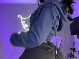 Pamelavegax poses in a blue hoodie and black leggings, showing her side profile and long dark hair against a purple lit background.