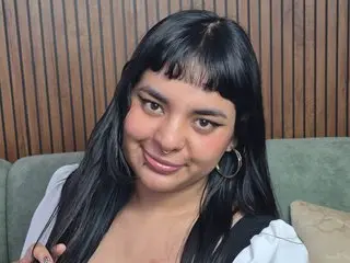 PastelWaifu with long black hair and bangs, wearing a white top and looking directly at the camera in front of a wood-paneled wall.