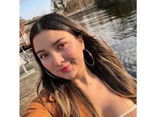 PaulinaSalaz poses with a smile, showing her long brown hair and cleavage while wearing an orange top outdoors.