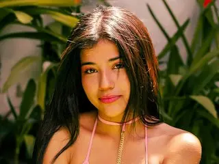 Peachrougeh poses with her long dark hair down, wearing a pink choker and thin pink straps against a background of green tropical leaves.
