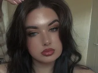 PeachyDoll23x looks directly at the camera with long, dark wavy hair and soft makeup.