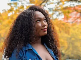 PearlBoller poses outdoors with her long, curly dark hair, wearing a blue button-down shirt against a soft-focus background of autumn leaves.