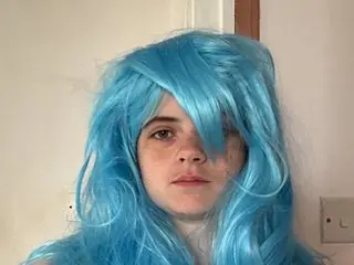 PearlPerry has long, bright blue hair and a neutral expression, looking directly at the camera against a plain white wall.