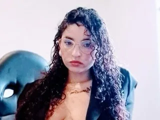 Penelope_Cox wears glasses and a black blazer while looking into the camera with her long curly hair down.