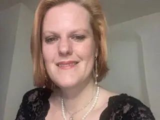 PennyGreen has red hair and wears a black lace top and a pearl necklace while posing against a plain white wall.