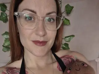 PerkyKaiaUK has long red hair and a light smile, wearing glasses and a black strap top while looking directly at the camera.