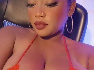 Perla_sexxy poses in a red bikini top, showing her cleavage and face with long eyelashes and gold hoop earrings.