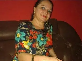 Perolasedutora smiles while sitting on a brown leather sofa, wearing a colorful floral print blouse with a gold necklace.